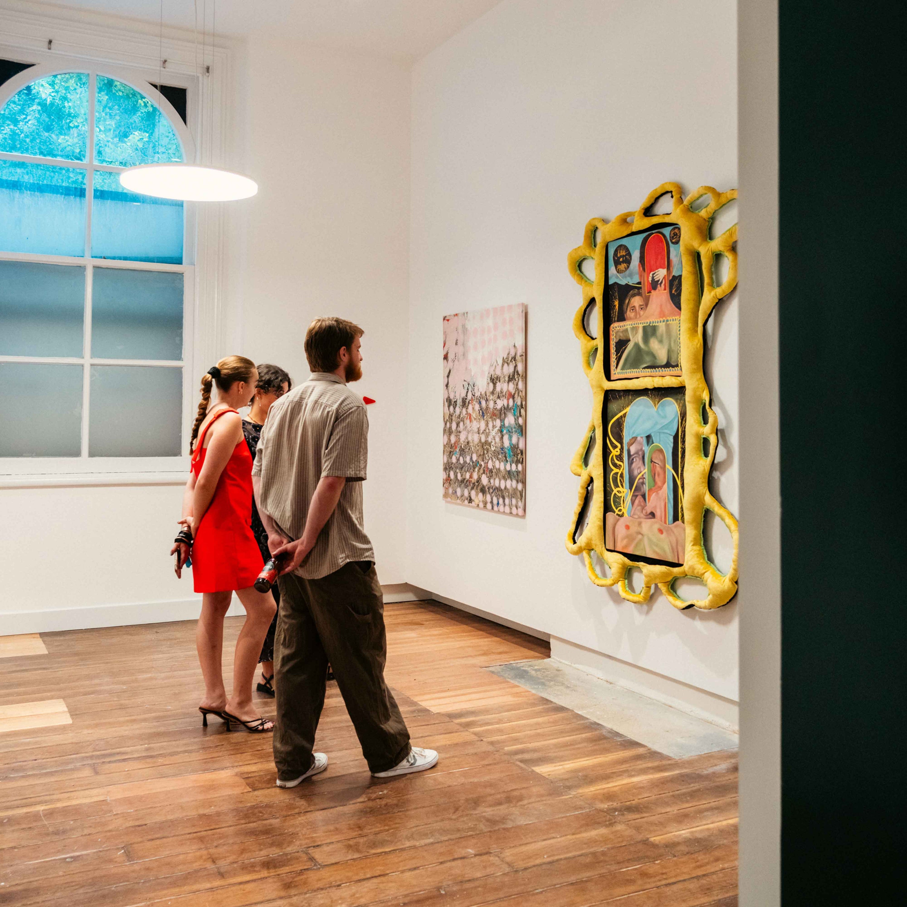 A white walled gallery displaying two colourful artworks on its walls. People are gathered in the middle as they view the artworks.