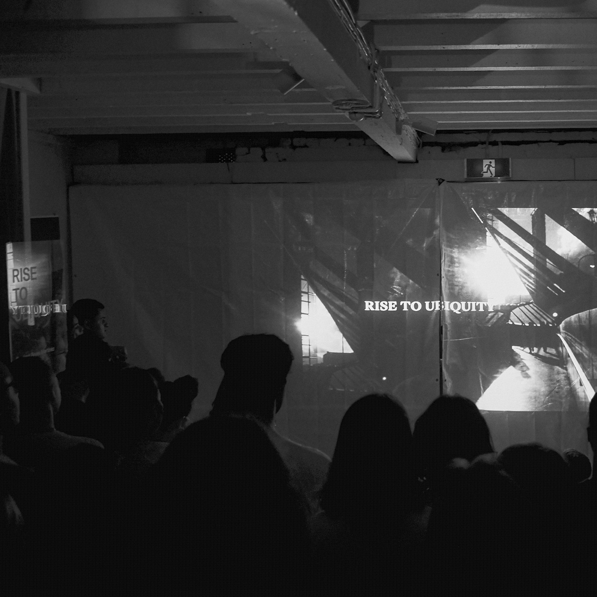 Black and white photo of Rowland Reyes Martinez's solo exhibition 'Rise To Ubiquity.' A large crowd watches the video installation for the short film projected on the gallery walls.