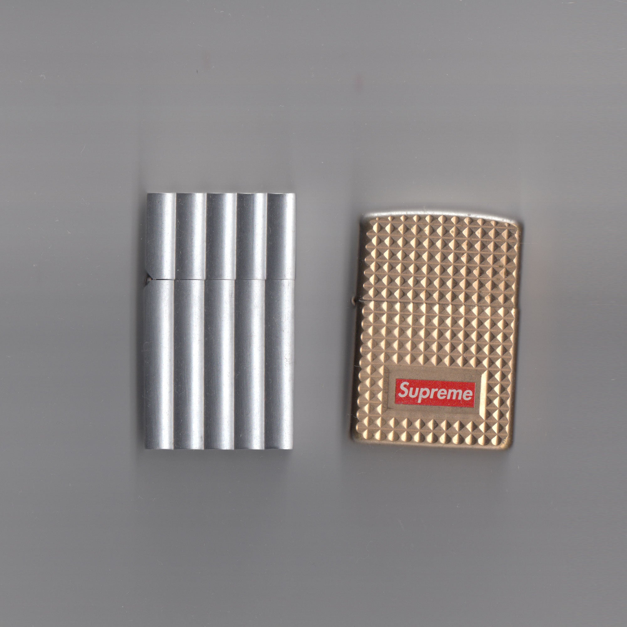 Scanned image of two lighters: one in grey stainless steel and the other gold, embellished with the red "Supreme" box logo imprint.